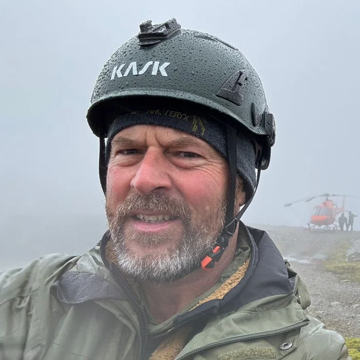 Bruce Shields wearing a safety helmet on location, with a helicopter behind him